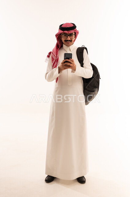 متابعة اّخر مستجدات العالم، استخدام التقنيات الحديثة والتكنولوجيا، بورتريه لطالب جامعي عربي خليجي سعودي مبتسم يرتدي الزي التقليدي يحمل حقيبة على ظهره ويمسك بيديه الهاتف المحمول يقوم بالتواصل مع الاهل والاقارب، الجسم بالطول الكامل، خلفية بيضاء