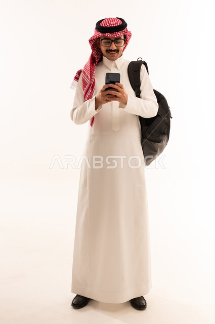 البحث والمطالعة من خلال الجوال, استخدام الأجهزة التقنية الحديثة, التعليم الجامعي في السعودية, بورتريه لطالب جامعي عربي خليجي سعودي مبتسم يرتدي الزي التقليدي يحمل حقيبة على ظهره ويمسك بيديه الهاتف المحمول, الجسم بالطول الكامل ، خلفية بيضاء