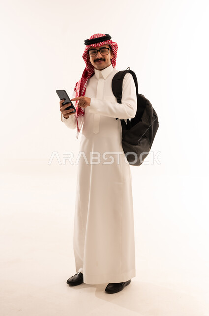 النظر إلى الكاميرا بإيماءات السعادة, استخدام الأجهزة التقنية الحديثة, التعليم الجامعي في السعودية, بورتريه لطالب جامعي عربي خليجي سعودي يرتدي الزي التقليدي يحمل حقيبة على ظهره ويمسك بيديه الهاتف المحمول, الجسم بالطول الكامل ، خلفية بيضاء
