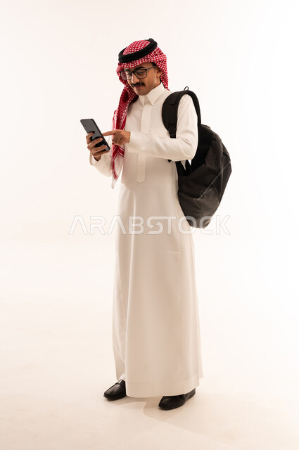 الاندماج والتمعن خلال التواصل مع الاصدقاء والاهل عبر الهاتف المحمول، ايماءات السعادة والسرور، استخدام التقنيات الحديثة والتكنولوجيا، بورتريه لطالب عربي خليجي جامعي يرتدي الثوب التقليدي يحمل حقيبه على ظهره ، الجسم بالطول الكامل، خلفية بيضاء