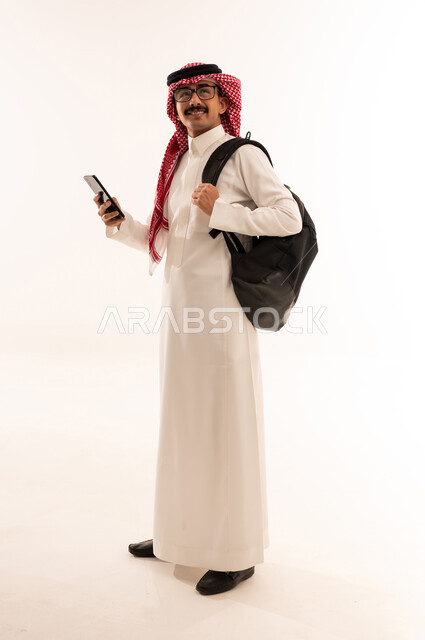 تصفح وسائل التواصل الاجتماعي عبر الجوال، استخدام الأجهزة التقنية الحديثة , بورتريه لطالب عربي خليجي سعودي مبتسم يرتدي الثوب التقليدي و الشماغ يحمل شنطة و يمسك الهاتف المحمول بيده ويقوم بالنظر الى شيء ما, صورة للجسم بالطول الكامل, خلفية بيضاء