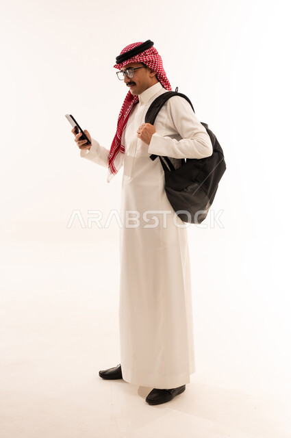 استخدام الأجهزة التقنية الحديثة , تصفح وسائل التواصل الاجتماعي عبر الجوال, بورتريه لطالب عربي خليجي سعودي مبتسم يرتدي الثوب التقليدي و الشماغ  يحمل شنطة يمسك الهاتف المحمول بيده, صورة للجسم بالطول الكامل, خلفية بيضاء