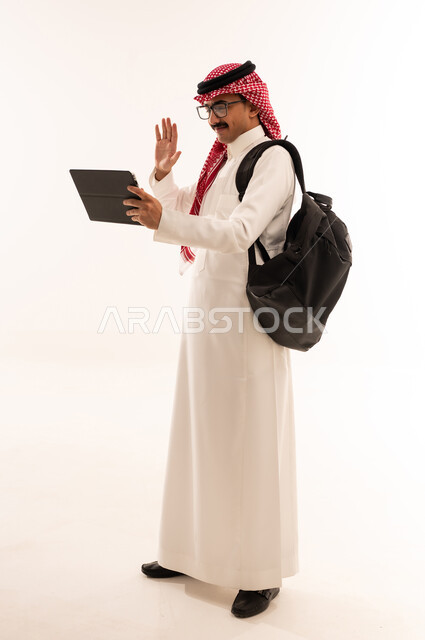 إستخدام أجهزه تقنية حديثة ، إيماءات تدل على السعادة ، بورتريه لطالب جامعي عربي خليجي سعودي مبتسم يرتدي الزي التقليدي يقوم بإجراء مكالمة فيديو من خلال التابلت ، الإستعداد للمحاضرات الجامعية الالكترونية ، التعلم عن بعد ، الجسم بالطول الكامل، خلفية بيضاء