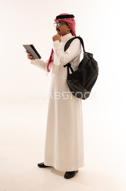 الإستعداد للمحاضرات الالكترونية، دمج التقنيات الحديثة بالتعليم الأكاديمي، بورتريه لطالب عربي خليجي سعودي مبتسم يرتدي الزي التقليدي يحمل جهاز التابلت و يقوم بالتفكير في شيء ما ، التعلم عن بعد ، مراجعة الدروس اليومية، الجسم بالطول الكامل، خلفية بيضاء