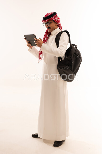 الإستعداد للمحاضرات الالكترونية ، إستخدام أجهزة تقنية حديثة ، بورتريه لطالب جامعي عربي يرتدي الزي الخليجي السعودي يقوم بإجراء مكالمة فيديو من خلال جهاز التابلت ، التعلم عن بعد ، مراجعة الدروس اليومية ، الجسم بالطول الكامل، خلفية بيضاء
