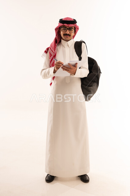 مراجعة الدروس اليومية، مفهوم الاجتهاد والتفوق، الاهتمام بالمذاكرة و الدروس ، التعليم الجامعي في السعودية, بورتريه لطالب جامعي عربي خليجي سعودي يرتدي  الزي التقليدي يحمل حقيبة على ظهره وبيده ورق ويقوم بدراسة المحاضرات, الجسم بالطول الكامل، خلفية بيضاء