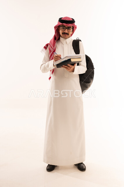مفهوم الاجتهاد والتفوق، استخراج المعلومات وتدوينها وكتابة التقارير والأبحاث العلمية, مراجعة الدروس اليومية, بورتريه لطالب جامعي عربي خليجي سعودي يرتدي الزي التقليدي يحمل حقيبة على ظهره وبيده كتاب ويقوم بدراسة المحاضرات, الجسم بالطول الكامل، خلفية بيضاء