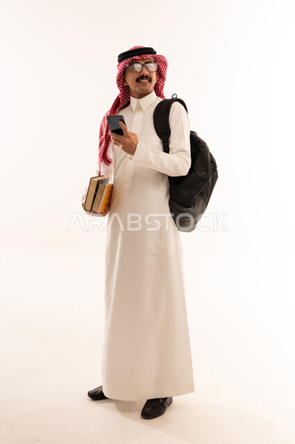 الاعجاب بالجامعة والمحاضرات، استخدام الأجهزة التقنية الحديثة, بورتريه لطالب جامعي عربي خليجي سعودي مبتسم يرتدي الزي التقليدي يحمل حقيبة على ظهره ويمسك بيديه الكتب والهاتف المحمول و ينظظر الى شيء ما, الجسم بالطول الكامل, خلفية بيضاء