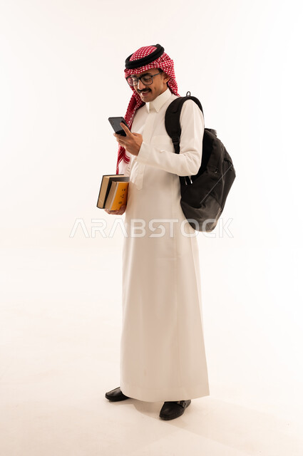 Following lectures and lessons, using modern technical devices, university education in Saudi Arabia, portrait of a smiling Saudi Arabian Gulf Arab university student wearing traditional clothing carrying a bag on his back and holding books and a mobile phone in his hands, full body, white background