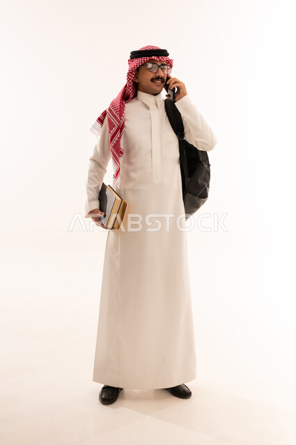 استخدام الأجهزة التقنية الحديثة, إجراء اتصال هاتفي عبر الجوال, التعليم الجامعي في السعودية, بورتريه لطالب جامعي عربي خليجي سعودي مبتسم يرتدي الزي التقليدي يحمل حقيبة على ظهره ويمسك بيديه الكتب والهاتف المحمول, الجسم بالطول الكامل, خلفية بيضاء