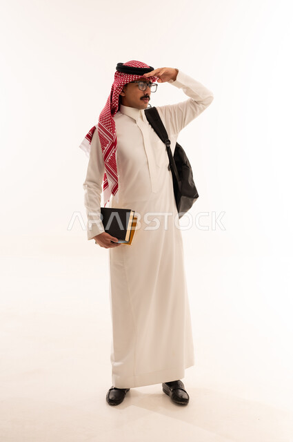 الاستعداد للعام الجامعي الجديد، الجامعات والكليات الأكاديمي، بورتريه لطالب عربي خليجي سعودي مبتسم يرتدي الزي السعودي التقليدي يحمل الحقيبة و الكتب الجامعية وينظرالى شيء ما، التعليم الدراسي في السعودية،الجسم بالطول الكامل، خلفية بيضاء