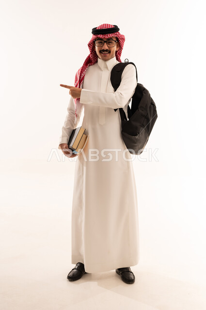 تطور التعليم في المملكه العربيه السعوديه، الاستعداد للعام الدراسي الجديد, بورتريه لطالب عربي خليجي سعودي مبتسم يرتدي الزي السعودي التقليدي يحمل الحقيبة و الكتب الجامعية و يشاور الى شيء ما، الجسم بالطول الكامل، خلفية بيضاء