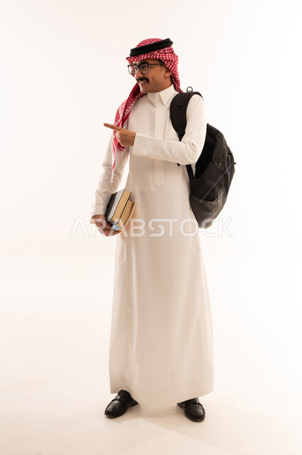 التعليم الدراسي في السعودية، الاستعداد للعام الدراسي الجديد, بورتريه لطالب عربي خليجي سعودي مبتسم يرتدي الزي السعودي التقليدي يحمل الحقيبة و الكتب الجامعية ينظر و يقوم بالاشارة الى شيء ما، الجسم بالطول الكامل، خلفية بيضاء