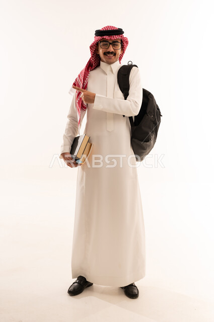 التحضير للذهاب إلى الجامعة، الاستعداد للعام الدراسي الجديد, بورتريه لطالب عربي خليجي سعودي مبتسم يرتدي الزي السعودي التقليدي يحمل الحقيبة و الكتب الجامعية و يشاور الى شيء ما، التعليم الدراسي في السعودية،الجسم بالطول الكامل، خلفية بيضاء