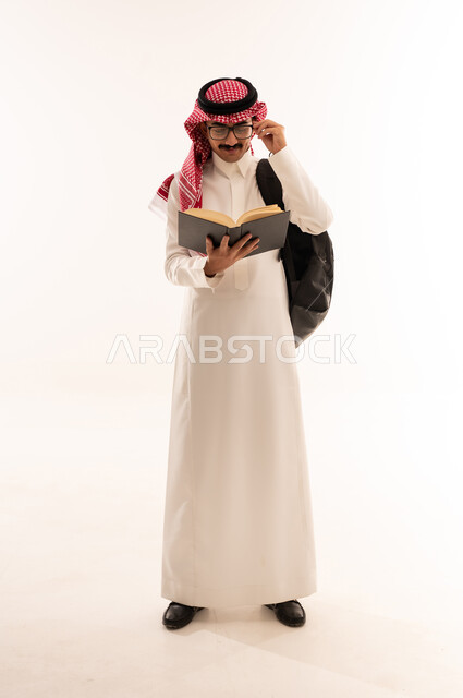 التعليم العالي في الجامعات والكليات الأكاديمية ، أهمية التعليم في المملكة العربية السعودية، بورتريه لطالب جامعي عربي خليجي سعودي يرتدي الزي التقليدي و النظارة والحقيبة الجامعية يقوم بقراءه الكتب الجامعية، الجسم بالطول الكامل، خلفية بيضاء