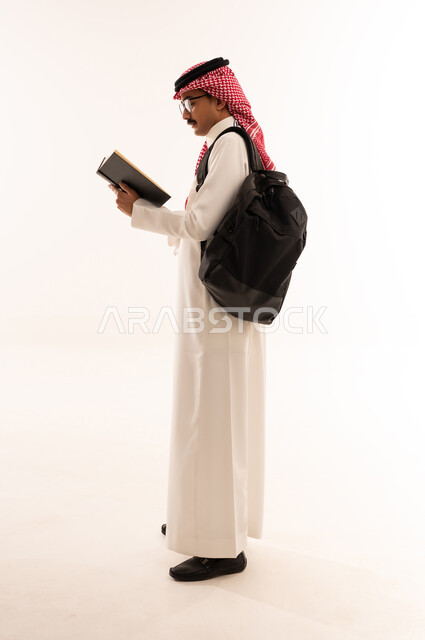 المناهج العلمية التعليمية، مراجعة الدروس اليومية ، أهمية التعليم في المملكة العربية السعودية، بورتريه من الجانب لطالب جامعي عربي خليجي سعودي يرتدي الزي التقليدي و النظارة والحقيبة الجامعية يقوم بقراءه الكتب الجامعية، الجسم بالطول الكامل، خلفية بيضاء