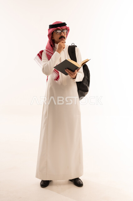 مراجعة الدروس اليومية ، إيماءات وجه ويدين تدل على الحيرة والتفكير، بورتريه طالب جامعي عربي خليجي سعودي يرتدي الزي التقليدي و نظارة يحمل بيده كتاب وعلى ظهره حقيبة ، التعليم الجامعي في المملكة العربية السعودية ،الجسم بالطول الكامل، خلفية بيضاء