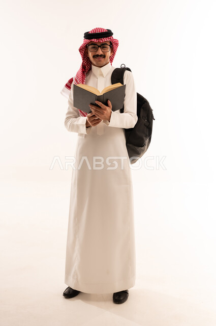 أهمية التعليم في المملكة العربية السعودية، بورتريه لطالب جامعي عربي خليجي سعودي مبتسم يرتدي الزي التقليدي و النظارة والحقيبة الجامعية يقوم بقراءه الكتب الجامعية، التعليم في المملكة العربية السعودية، الجسم بالطول الكامل، خلفية بيضاء