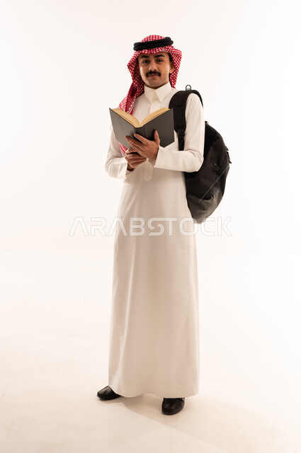 تطوير المناهج التعليمية في السعودية، الاجتهاد وتحضير الدروس, بورتريه لشاب عربي سعودي خليجي مبتسم يرتدي الثوب التقليدي يحمل الحقيبة يقوم بقراءة الكتب و ينظر الي شيء ما, بداية العام الدراسي الجديد, صورة الجسم بالكامل،الجسم بالطول الكامل، خلفية بيضاء