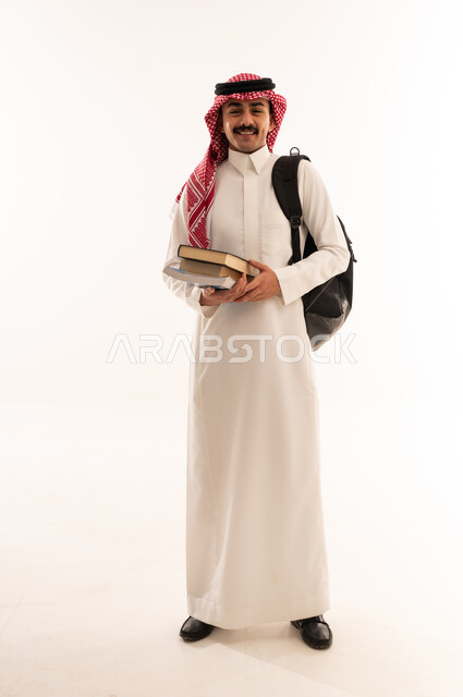 الاستعداد للعام الدراسي الجديد, بورتريه لطالب عربي خليجي سعودي مبتسم يرتدي الزي السعودي التقليدي يحمل الحقيبة وبيده الكتب الجامعية، الإستعداد للعام الدراسي الجديد، التعليم الدراسي في السعودية،الجسم بالطول الكامل، خلفية بيضاء