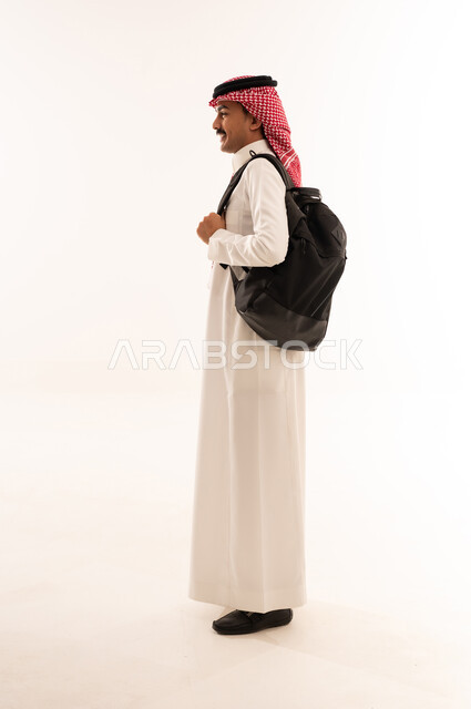 متابعة المحاضرات والدروس اليومية ، الاستعداد للذهاب الى الدوام الجامعي، التعليم في المملكة العربية السعودية، بورتريه من الجانب لطالب جامعي عربي خليجي سعودي مبتسم يرتدي الزي التقليدي والحقيبة الجامعية، الجسم بالطول الكامل، خلفية بيضاء