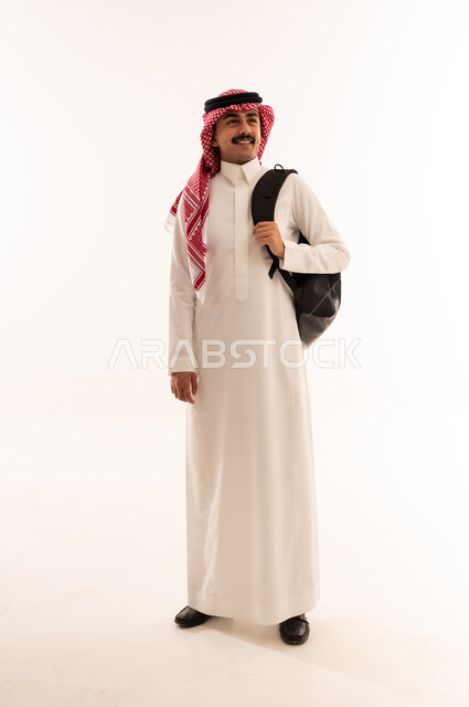 الاستعداد للذهاب الى الدوام الجامعي، التعليم في المملكة العربية السعودية، بورتريه لطالب جامعي عربي خليجي سعودي مبتسم يرتدي الزي التقليدي والحقيبة الجامعية ينظر الي شي ما، متابعة المحاضرات والدروس اليومية ،الجسم بالطول الكامل، خلفية بيضاء