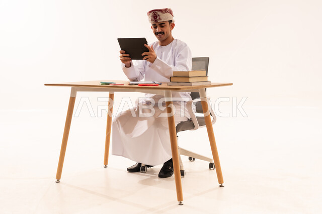 مفهوم التعليم عن بعد، البحث الالكتروني عبر الانترنت, استخدام الأجهزة والتقنيات الحديثة والمتطورة، بورتريه لطالب جامعي عربي خليجي عماني مبتسم يرتدي الكمه والدشداشه يجلس على الطاولة ويمسك جهاز التابليت, مراجعة ومتابعة الدروس والواجبات اليومية, خلفية بيضاء