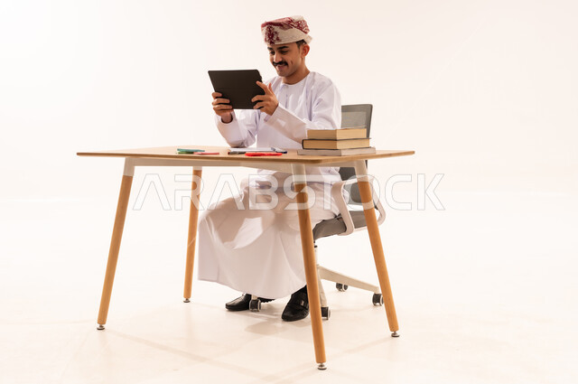 البحث الالكتروني عبر الانترنت, مفهوم التعليم عن بعد, استخدام الأجهزة والتقنيات الحديثة والمتطورة، بورتريه لطالب جامعي عربي خليجي عماني مبتسم يرتدي الكمه والدشداشه يجلس على الطاولة ويمسك جهاز التابليت, مراجعة ومتابعة الدروس والواجبات اليومية, خلفية بيضاء
