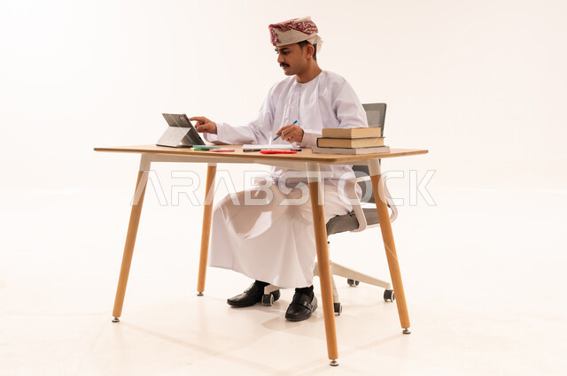 البحث الالكتروني عبر الانترنت, استخدام الأجهزة والتقنيات الحديثة والمتطورة, مفهوم التعليم عن بعد, بورتريه لطالب مقرب جامعي عربي خليجي عماني يرتدي الكمه والدشداشه يجلس على الطاولة ويعمل على جهاز التابليت, مراجعة ومتابعة الدروس والواجبات اليومية, خلفية بيضاء