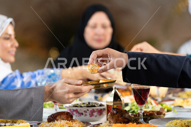 A table full of food and drinks, Islamic and Ramadan rituals, family discussions and dialogues at the dining table, spending enjoyable family times, a close-up of the hand of an Arab Gulf Emirati woman presenting a plate of oriental sweets, gestures of happiness and pleasure