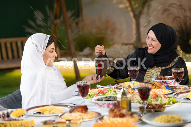 أجواء عائلية دينية رمضانية ، تبادل الأحاديث بين أفراد الأسرة، سفرة مليئة بأصناف من المأكولات الشرقية الشهية، أداء فريضة الصيام في شهر رمضان المبارك، امراة عربية خليجيه اماراتية تقوم بتقديم الفانوس لابنتها بايماءات السعاده و السرور