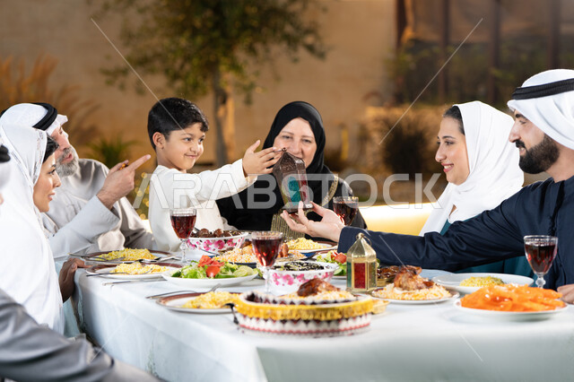 اجتماع الاسرة حول مائدة الطعام، نقاشات وحوارات عائلية، مائدة طعام مليئة بالوجبات الرمضانية المتنوعة، الإستمتاع  باجواء رمضان مع الأهل، أجواء أسرية مليئة بالحب، ولد عربى خليجي اماراتي يقوم بتقديم فانوس رمضان لوالده بايماءات السعاده و السرور