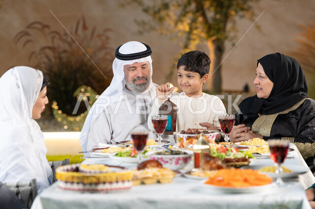 أجواء شهر رمضان المبارك، مائدة طعام مليئة بالوجبات الرمضانية المتنوعة، نقاشات وحوارات عائلية، أجواء أسرية مليئة بالحب، اجتماع الاسرة حول مائدة الطعام، ولد عربى خليجي اماراتي يقوم بامساك فانوس رمضان بايماءات السعاده و السرور