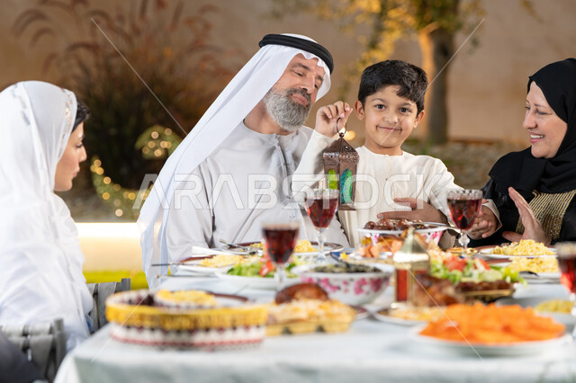 الأعياد والمناسبات الاسلامية،  مائدة طعام مليئة بالوجبات الرمضانية المتنوعة، نقاشات وحوارات عائلية، أجواء أسرية مليئة بالحب، اجتماع الاسرة حول مائدة الطعام، ولد عربى خليجي اماراتي يقوم بامساك فانوس رمضان بايماءات السعاده و السرور