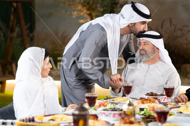 أجواء عائلية رمضانيه, سفرة إفطار مليئة بأصناف من المأكولات والمقبلات الشهية, عائلة عربية خليجية إماراتية مستمتعة بتناول الفطور في شهر رمضان, إجتماع الأهل على مائدة الافطار, مشاركة الطعام وإكرام الوالدين، شاب اماراتي خليجي  يقوم بتقبيل رأس والده، التربية الصالحة للأبناء