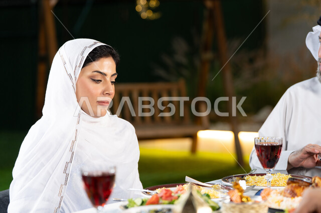 حلول موعد الإفطار عند اذان المغرب، سفرة مليئة بأصناف المأكولات والمقبلات الشهية، الوجبات والأطباق الرئيسية على مائدة الطعام، صورة مقربة لامراة عربيه خليجيه اماراتية ترتدي الحجاب و تقوم بالنظر الى المائده