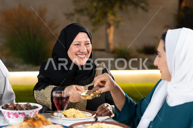 حسن الضيافة والاستقبال في شهر رمضان المبارك، سفرة مليئة بأصناف من المأكولات الشرقية الشهية، أجواء عائلية دينية رمضانية سعيدة، امراة عربية خليجية اماراتية مبتسمه تقوم بتقديم الحلوي
