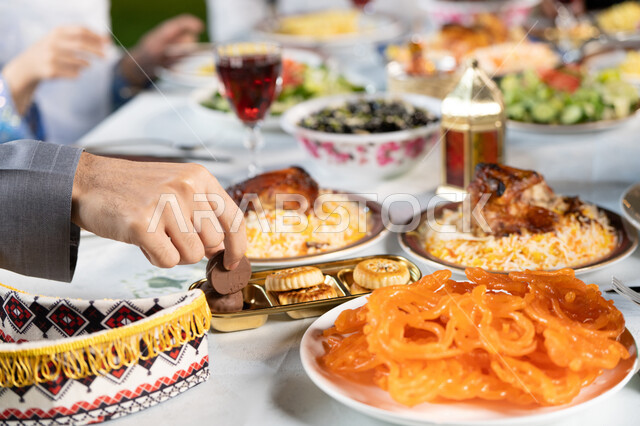 حسن الضيافة والاستقبال في شهر رمضان المبارك، صورة مقربة ليد رجل عربي يقوم بتناول  حلوي اللقيمات الشهية، الأجواء الرمضانية الروحانية، عائله عربية خليجيه اماراتية تجتمع حول الطعام، سفرة مليئة بأصناف المأكولات والمقبلات الشهية