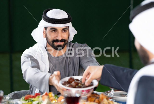 أجواء رمضانية روحانية،  الكرم والمشاركة خلال وقت الإفطار، جلسة عائلية مليئة بالمحبة والاحترام، صورة مقربة لرجل خليجي إماراتي يقدم طبق التمر لأحد أفراد العائلة، ايماءات السعادة و السرور بقدوم شهر رمضان المبارك