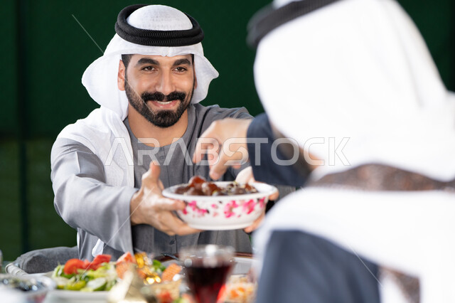 الكرم والمشاركة خلال وقت الإفطار، جلسة عائلية مليئة بالمحبة والاحترام, أجواء رمضانية روحانية، صورة مقربة لرجل خليجي إماراتي يقدم طبق التمر لأحد أفراد العائلة، تجمع العائلة على سفرة الاكل، ايماءات السعادة و السرور بقدوم شهر رمضان المبارك