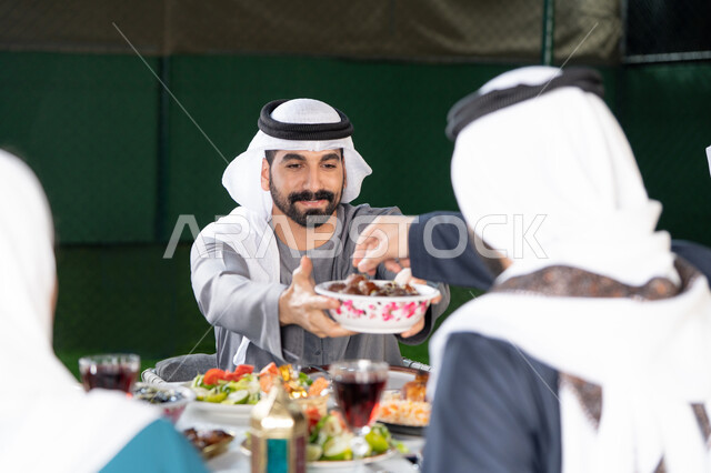 الكرم والمشاركة خلال وقت الإفطار, أجواء رمضانية روحانية, جلسة عائلية مليئة بالمحبة والاحترام، رجل خليجي إماراتي يقدم طبق التمر لأحد أفراد العائلة، تجمع العائلة على سفرة الاكل، ايماءات السعادة و السرور بقدوم شهر رمضان المبارك