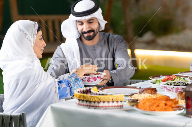 قضاء أوقات ممتعة مع  الاسره في الهواء الطلق، مائدة إفطار مليئة بالأصناف والمأكولات الشهية،  زوجان عربيان خليجيان اماراتيان يقومان بتناول الافطار بايماءات السعادة و السرور، رجل خليجي يقوم بتقديم طبق التمر لزوجته، الشهر الكريم والأجواء الرمضانية الروحانية