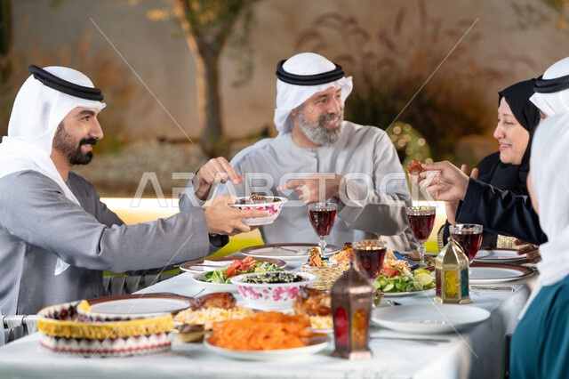حسن الضيافه والإستقبال, إفطار صائم، قضاء أوقات ممتعة مع العائلة في الهواء الطلق، مائدة إفطار مليئة بالأصناف والمأكولات الشهية، عائلة عربية خليجية إماراتية تقوم بتناول الافطار بايماءات السعادة و السرور، رجل خليجي يقوم بتقديم طبق التمر للضيوف