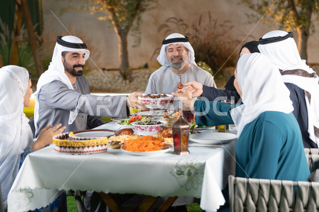 The holy month and the spiritual Ramadan atmosphere, spending enjoyable times with the family outdoors, an Iftar table filled with delicious dishes and foods, an Arab Gulf Emirati family eating dates with gestures of happiness and pleasure