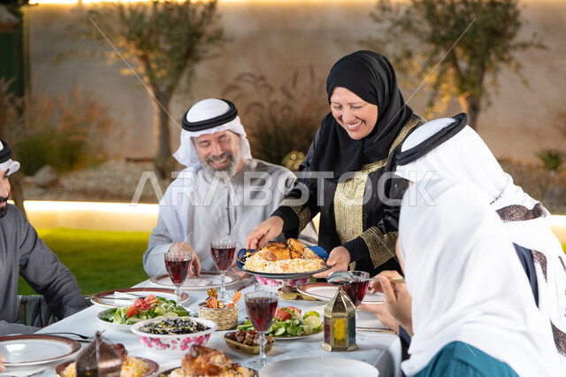 تجمع العائلة على سفرة الاكل، الاحتفال بالاعياد و المناسبات الاسلامية, امراة تقوم بسكب الطعام في الصحون و توزيعه على افراد الاسرة, ايماءات السعادة و السرور بقدوم شهر رمضان المبارك, عائلة عربية خليجية اماراتية تجلس على الطاولة المليئة بالاطباق الشهية