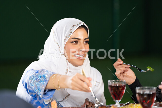 اجواء رمضانية روحانية، تقديم المأكولات على مائدة الإفطار,صورة مقربة  لامرأة عربية خليجية اماراتية مبتسمه ترتدي الثوب التقليدي و الحجاب تقوم بتناول الطعام , أجواء شهر رمضان المبارك في الامارات, المناسبات الاسلامية و الدينية