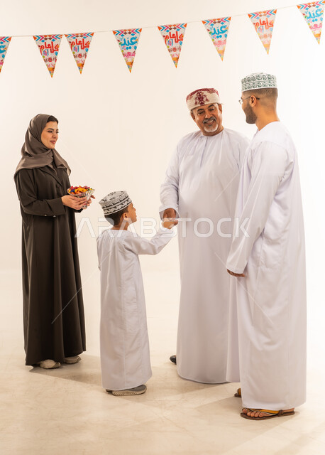 إظهار مشاعر المودة والمحبة العائلية, أجواء البهجة والسعادة في العيد, بورتريه لعائلة عربية خليجية عمانية تحتفل بالأعياد الاسلامية, الديكورات والزينة الرمضانية ،التحية والمصافحة بين أفراد الأسرة والتهنئات, تقديم الحلويات التقليدية والمأكولات الشهية, خلفية بيضاء .