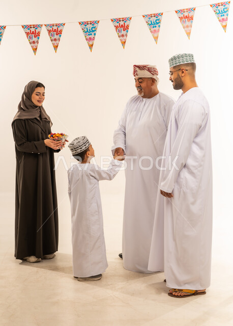 أجواء البهجة والسعادة في العيد, بورتريه لعائلة عربية خليجية عمانية تحتفل بالأعياد الاسلامية, التحية والمصافحة بين أفراد الأسرة و التهنئات, الديكورات والزينة الرمضانية ، تقديم الحلويات التقليدية والمأكولات الشهية, إظهار مشاعر المودة والمحبة العائلية, خلفية بيضاء .