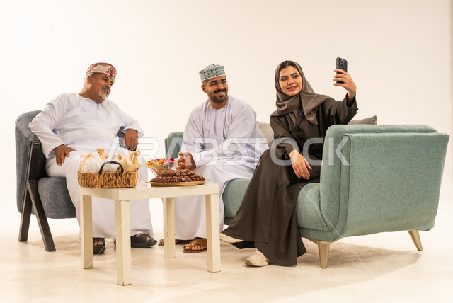 Offering Arabic hospitality and traditional sweets, taking souvenir photos with a mobile phone, a portrait of an Arab Gulf Omani family wearing traditional costumes celebrating the coming of Eid, the concept of love and affection, sitting in the living room in a family atmosphere and exchanging greetings, white background.
