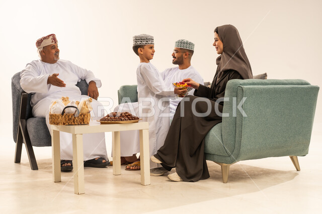 مشاركة اجواء رائعة لعائلة عمانية داخل المنزل تحتفل بالعيد ، زينة وديكورات الأعياد ، أجواء العيد في المجتمع العماني ، الزيارات العائلية وتجمعات الأسر ، ابن يقدم الضيافة والحلوى لوالدته ، بورتريه لعائلة عربية خليجية عمانية تحتفل بالأعياد , خلفية بيضاء .